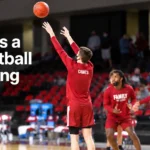 Basketball player taking a warm-up shot while wearing a red long-sleeve basketball shooting shirt, with teammates warming up in the background.